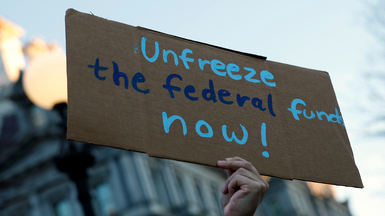 A hand holds a cardboard sign up that reads "unfreeze the federal funds now!" in blue lettering.