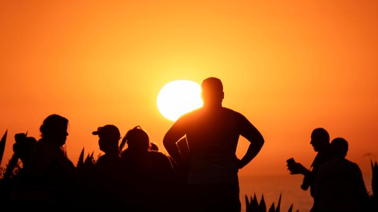 Six people are silhouetted against an orange sunset, with the person in the center standing with their hands on their hips